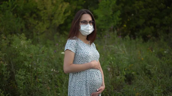 Mladá těhotná žena v lékařské masce stojí v přírodě. Dívka v brýlích a šatech se dívá do kamery. — Stock fotografie