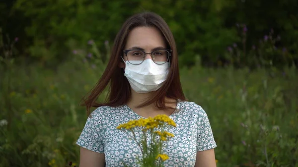 Egy fiatal nő orvosi maszkban virágot szimatol és rázza a fejét, mert nincs szaga. Szemüveges lány és ruha a természetben. — Stock Fotó