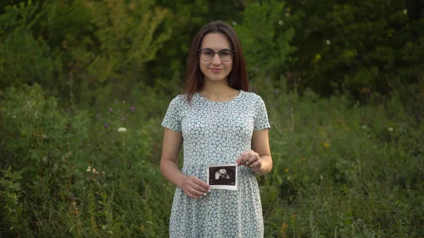 O tânără gravidă deține o imagine cu ultrasunete a unui copil în mâini. Fata în ochelari și o rochie în natură . — Fotografie, imagine de stoc