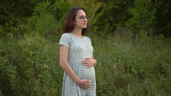 Uma jovem grávida levanta-se e segura a barriga com as mãos. Menina em óculos e um vestido na natureza. — Fotografia de Stock