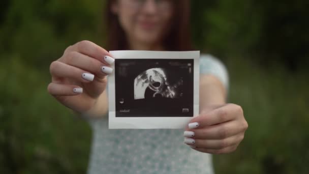 Een jonge zwangere vrouw houdt een foto van een baby in haar handen. Meisje in de natuur. — Stockvideo