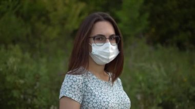 Tıbbi maskeli hamile bir kadın doğada duruyor. Gözlüklü ve elbiseli kız kameraya bakıyor..