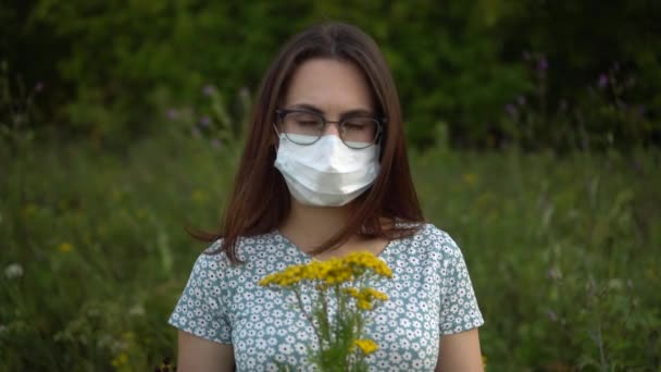 Uma jovem com uma máscara médica cheira flores e balança a cabeça porque não cheira mal. Menina em óculos e um vestido na natureza. — Vídeo de Stock