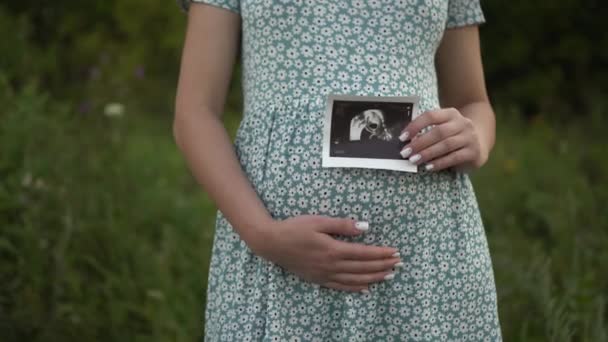 Una giovane donna incinta tiene in mano un'immagine ecografica di un bambino. Ragazza in un vestito all'aperto. — Video Stock