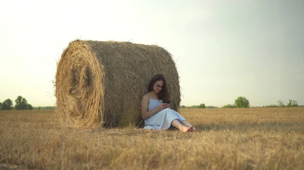 Una giovane donna siede vicino a un pagliaio in un campo con un telefono in mano. Una ragazza con gli occhiali e un vestito blu sta messaggiando al telefono. — Foto Stock