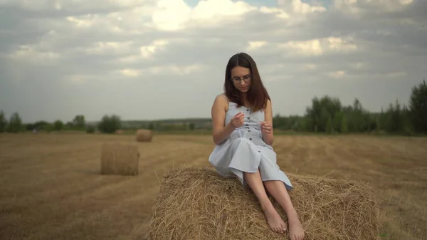 Die junge Frau sitzt auf einem Heuhaufen auf einem Feld. Ein Mädchen mit Brille und blauem Kleid. — Stockfoto