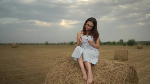 Una joven se sienta en un pajar en un campo. Una chica con gafas y un vestido azul. — Foto de Stock