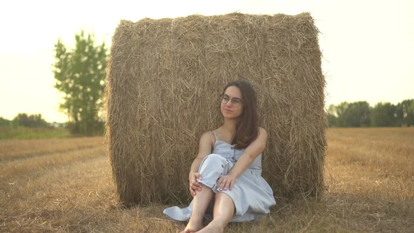 Eine junge Frau sitzt in der Nähe eines Heuhaufens auf einem Feld. Ein Mädchen mit Brille und blauem Kleid ruht auf dem Feld. — Stockfoto