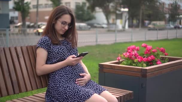 Jeune fille enceinte avec un téléphone dans ses mains. Une fille avec des lunettes et une robe envoie des textos au téléphone. — Video