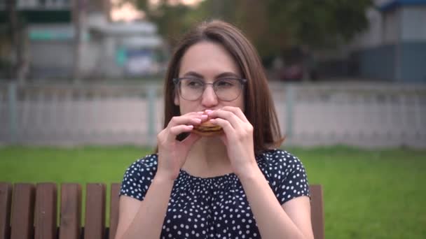 Młoda ciężarna dziewczyna je hamburgera siedząc na ławce w parku. Dziewczyna w okularach i sukience. Kamera się odsuwa.. — Wideo stockowe