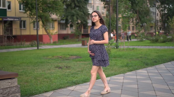Une jeune fille enceinte marche le long de l'allée et touche son estomac avec ses mains. Fille avec des lunettes et une robe dans le parc. — Photo