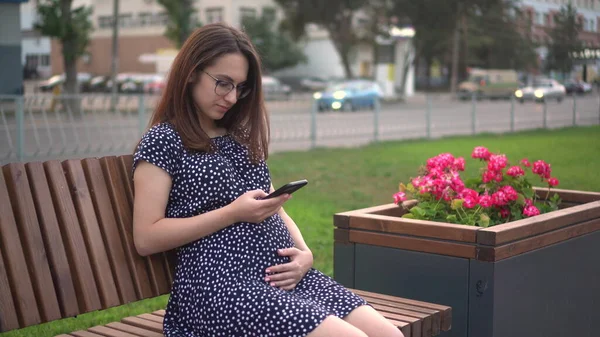 Joven embarazada con un teléfono en las manos. Una chica con gafas y un vestido está enviando mensajes por teléfono. — Foto de Stock