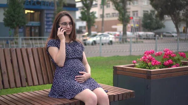 Молодая беременная девушка разговаривает по телефону в парке. Девушка в очках и платье. — стоковое фото