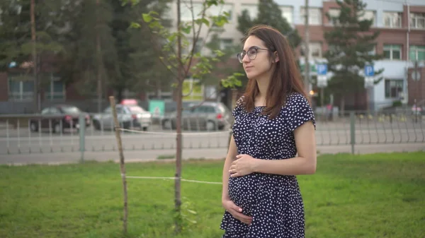 Молодая беременная девушка идет по переулку и прикасается руками к животу. Девушка в очках и платье в парке. — стоковое фото