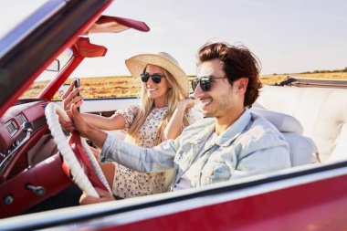 Bir kadın erkek arkadaşıyla araba gezisinde selfie çekiyor.                               