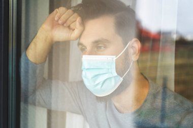 Resigned man with mask looking out of window                                clipart