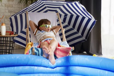 Güneş gözlüklü rahat çocuk evde bir sandalyede uzanıyor.