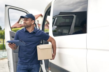 Paketi olan bir kurye teslimat arabasının yanına bakar.