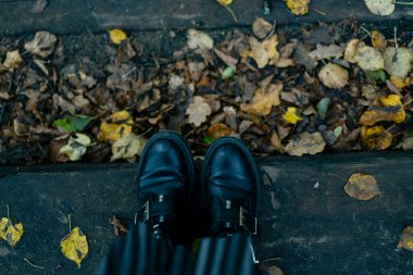 Sonbahar yapraklarıyla kaplı tahtaların üzerinde duran siyah deri çizmeler giyen biri. Sıcak bir sonbahar atmosferi, kırsal bir hava ve sarı yapraklar.