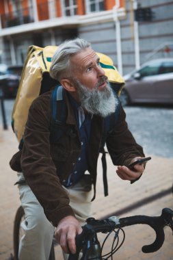Yaşlı sakallı bir adamın dikey resmi. Lezzetli bir adam olarak çalışıyor, bisiklete biniyor, akıllı telefon kullanıyor.