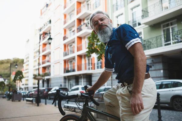 Yaşlı, sakallı, gri saçlı bir adamın kameraya gülümserkenki düşük açılı görüntüsü.