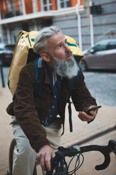 Yaşlı sakallı bir adamın dikey resmi. Lezzetli bir adam olarak çalışıyor, bisiklete biniyor, akıllı telefon kullanıyor.