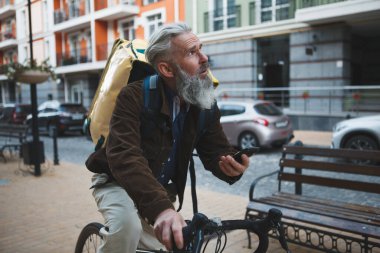 Teslimat servisinde çalışan yaşlı bir adam, adresi bulmak için akıllı telefon kullanıyor, bisiklete biniyor.