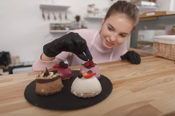 Cheerful female confectioner decorating desserts with fruits and berries