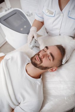 Kozmetik uzmanı tarafından profesyonel cilt bakımı gören sakallı yakışıklı bir adamın dikey çekimi.