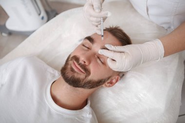 Genç adam, kozmetolojist tarafından kırışıklık önleyici tedavi için enjeksiyon yaptırıyor.