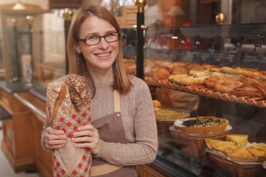 Kameraya gülümseyen ve elinde bir somun taze ekmek tutan olgun kadın fırıncı.