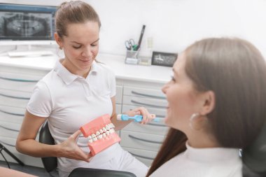 Kadın dişçi hastasına dişlerini nasıl fırçalayacağını gösteriyor.