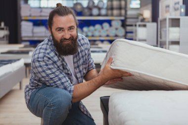 Sağlıklı uyku konsepti. Yakışıklı, sakallı, kameraya gülümseyen olgun bir adam. Ev eşyaları mağazasından yeni ortopedik yatak alıyor. Neşeli erkek müşteri yeni yatak seçiyor.