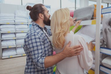 Arka planda, bir çiftin sarılma ve alışveriş yaparken çekilmiş görüntüleri var. Karı koca mobilya mağazasının raflarından ortopedik yastıklar seçiyor.