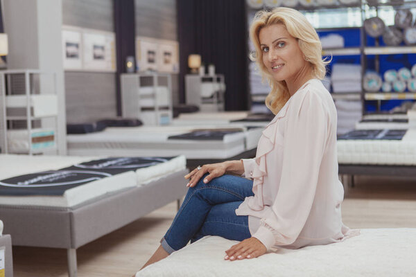 Очаровательная зрелая женщина, улыбающаяся в камеру, сидя на новой ортопедической кровати в мебельном магазине, копирует пространство. Счастливая женщина покупает удобный матрас для здорового сна
