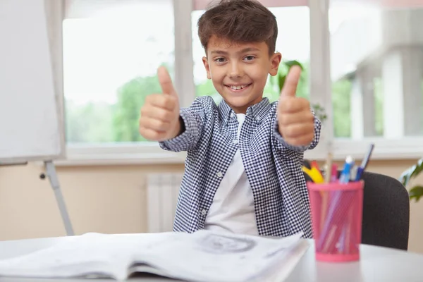 宿題をしながら嬉しそうに親指を立てて微笑む幸せな少年 勉強を楽しむチャーミングな少年 子供時代のコンセプト — ストック写真