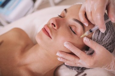 Güzellik kliniğinde gençleştirici yüz enjeksiyonları yaptıran güzel bir genç kadın. Kozmetik uzmanı bir müşterinin derisine yüz doldurucu enjekte ediyor.
