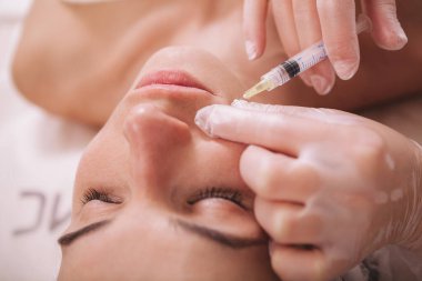 Güzeller güzeli genç bir kadının güzellik kliniğinde yüz dolgu iğnesi olurken çekilmiş bir fotoğrafı. Kozmetik uzmanı, tedavi seansında bir müşterinin cildine hyaluronik asit enjekte ediyor.