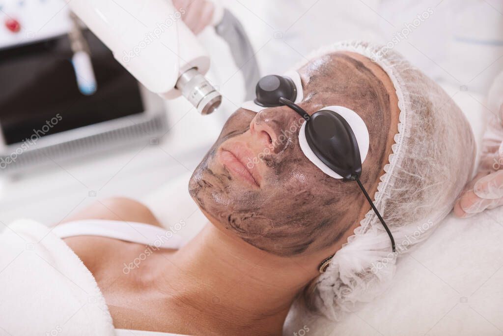 Close up of a mature woman wearing protective eyeglasses, getting carbon laser peel treatment by cosmetologist