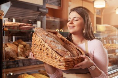 Güzel genç bayan girişimci fırınında çalışmaktan zevk alıyor, sepetteki lezzetli taze ekmek kokusunu alıyor.