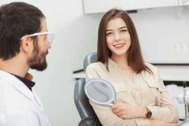 Kameraya gülümseyen güzel bir kadın, klinikte dişçiyi ziyaret ederken bir ayna tutuyor.