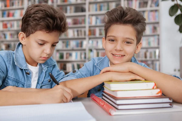 双子の弟が教科書に宿題を書いている間 カメラに笑みを浮かべているかわいい幸せな少年 学習の概念 — ストック写真