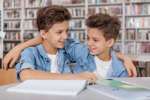 一緒に学校の宿題をしながら図書館で抱き合って笑っている幸せな若い双子の兄弟 — ストック写真