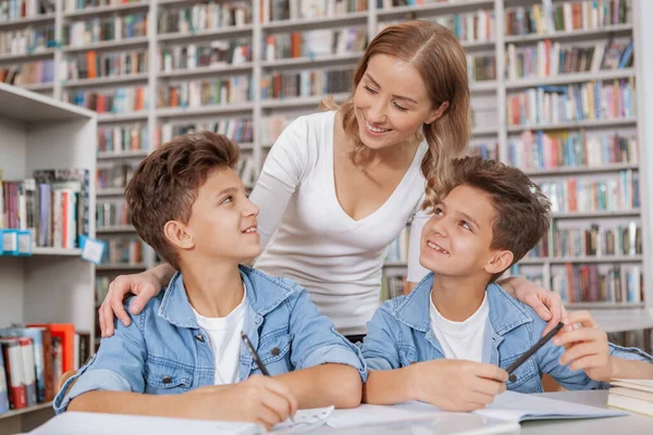 図書館で宿題をして子供たちを助ける魅力的な女性 双子の兄弟は一緒に勉強しながら母親に微笑んだ — ストック写真