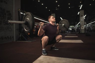 Genç, kaslı erkek sporcu, spor salonunda ağır halterle çömeliyor.