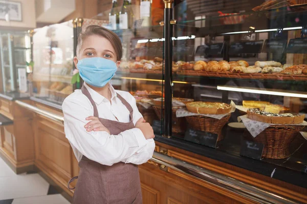 Young boy wearing medical face mask and apron, working at his family bakery shop, copy space