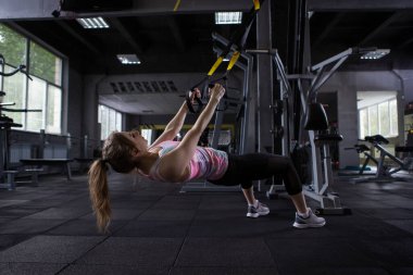 Spor salonundaki trx antrenörü üzerinde düşük sıra egzersizi yapan bayan sporcu.