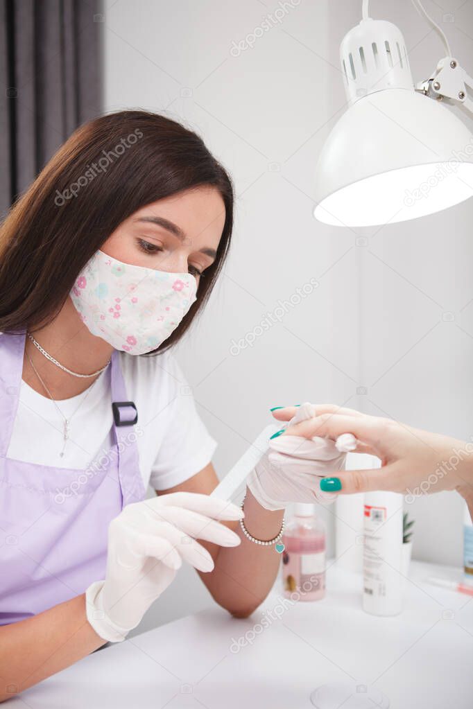 Vertical shot of female manicurist filing nails of client, wearing face mask