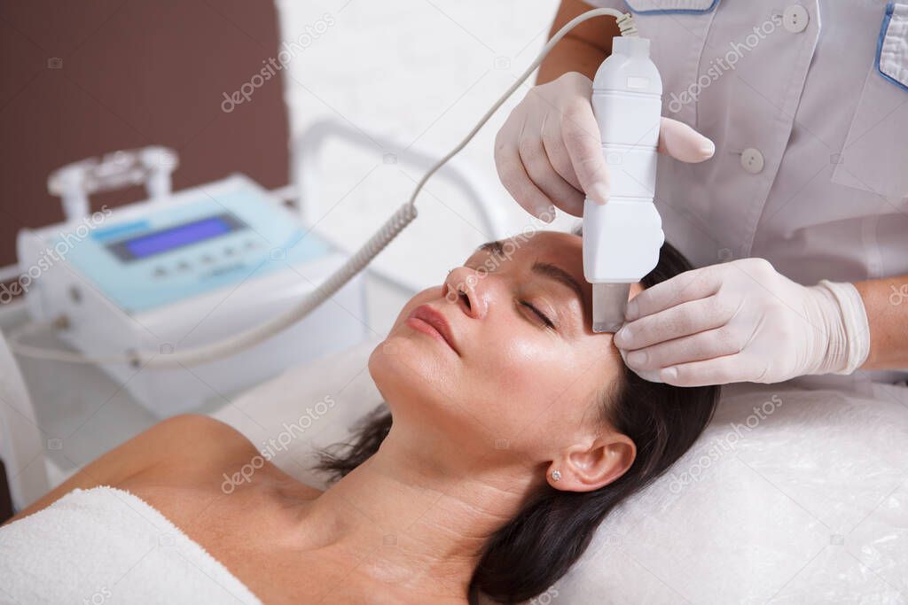 Unrecognizable cosmetologist doing ultrasound skincare facial for mature woman