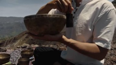 Tibedt çalan genç bir Asyalı doğadaki bir dağda bakır bardaklar söylüyor.